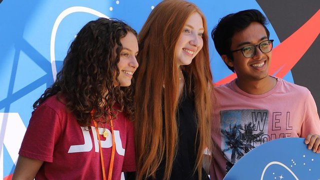 Three interns pose together in front of the NASA meatball holding JPL and NASA signs.